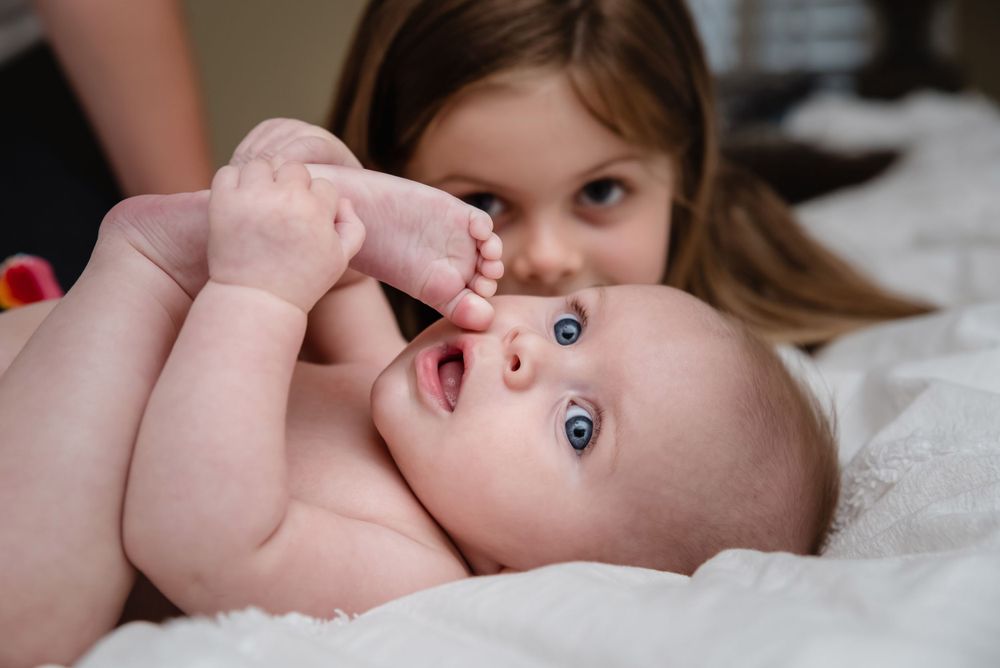 Blue-eyed baby lying on white blanket with older sister smiling behind.
