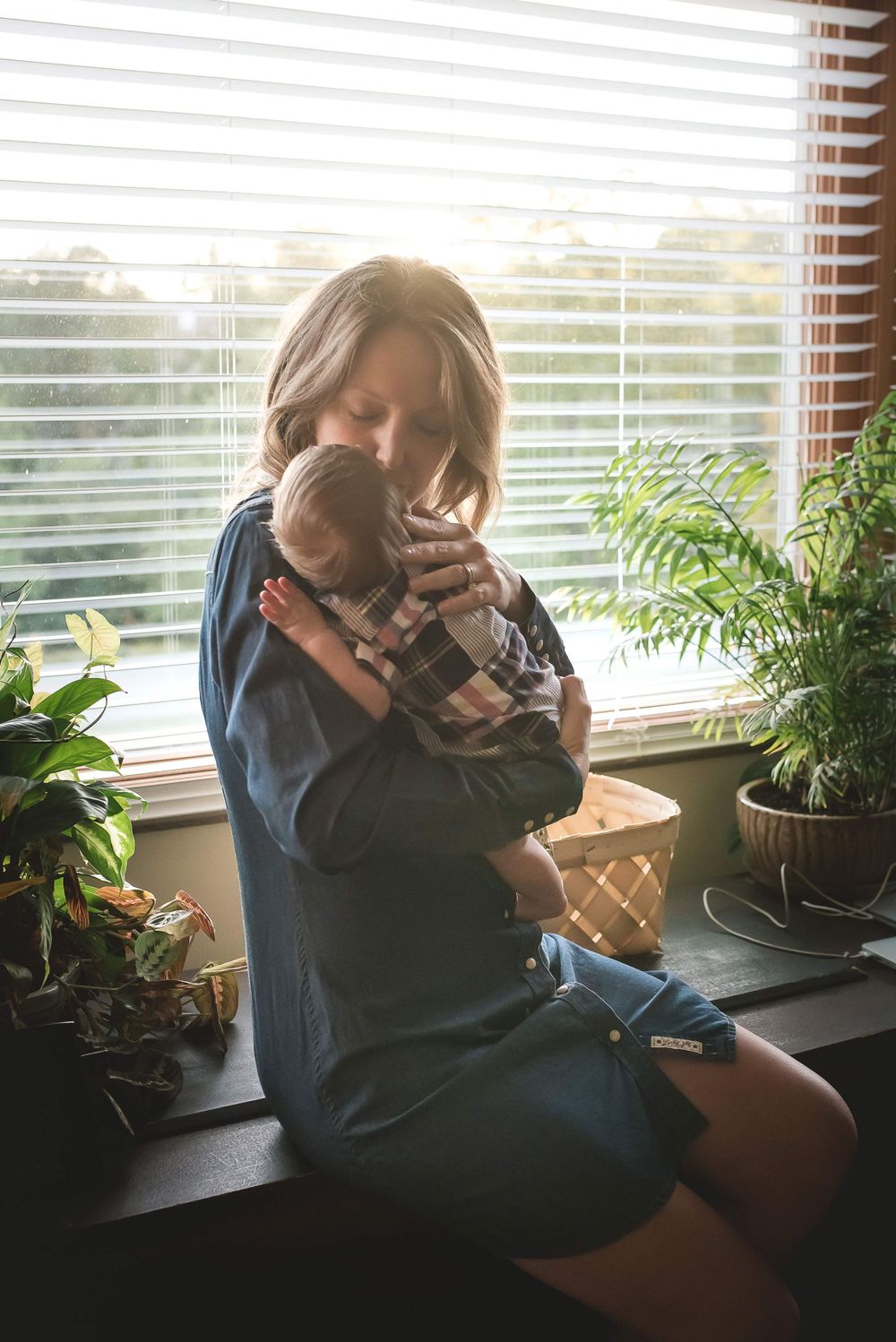 Mother holding newborn baby near sunlit window surrounded by plants.
