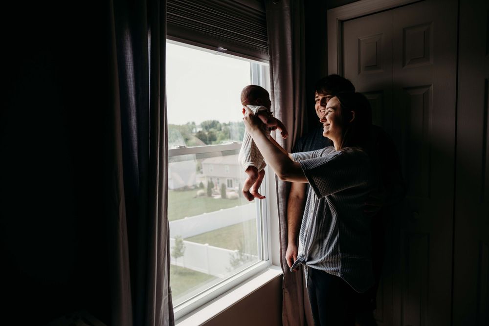 Parent holding newborn up to look out bright bedroom window.