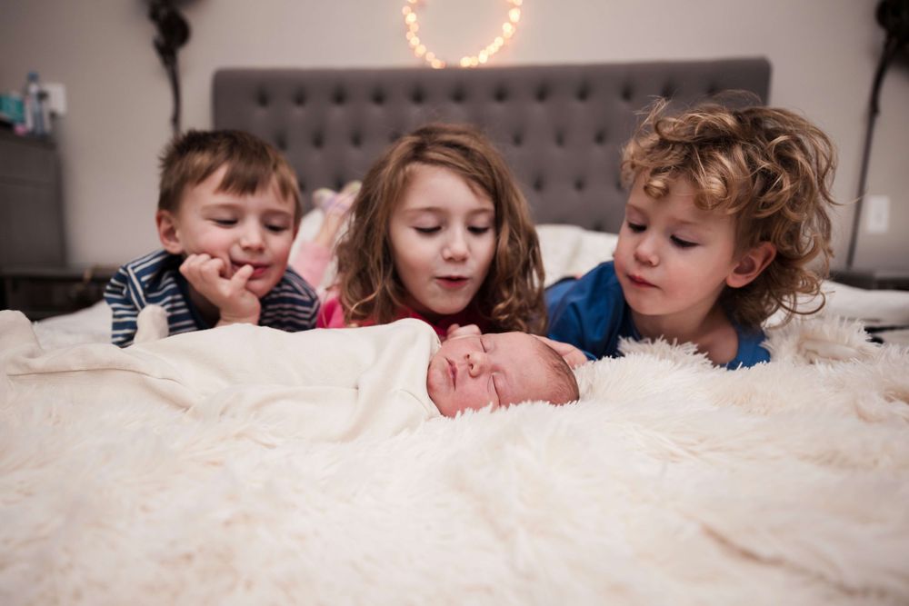 Three young siblings lying on white bed smiling at sleeping newborn baby.