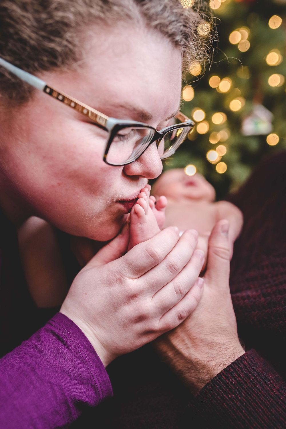 A woman wearing glasses kisses a newborn baby's tiny foot near a Christmas tree.