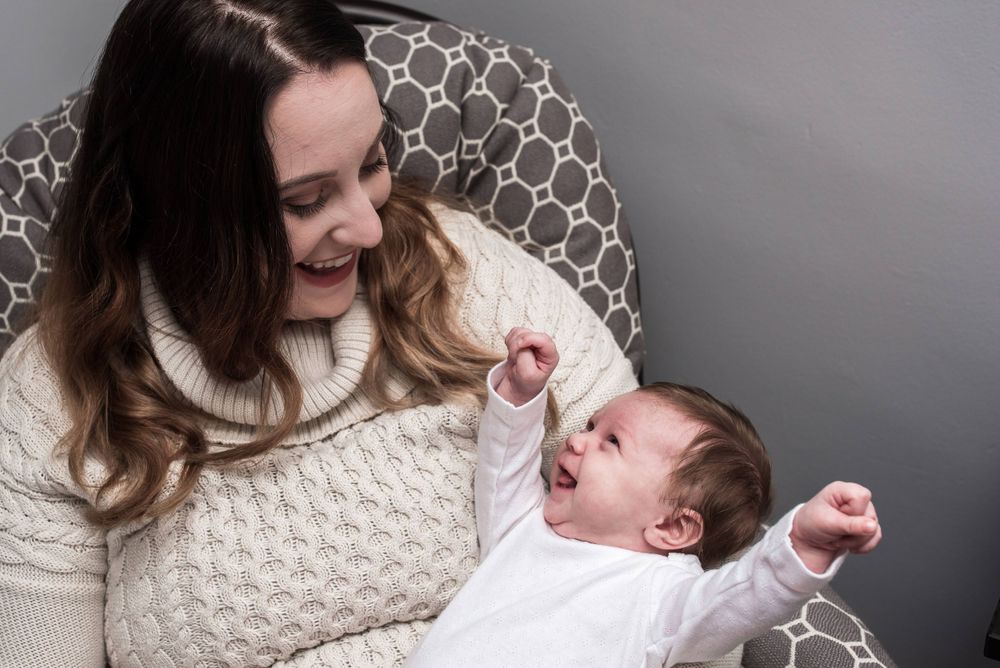 A smiling mother in a white sweater watches her happy baby stretch arms wide in a chair.