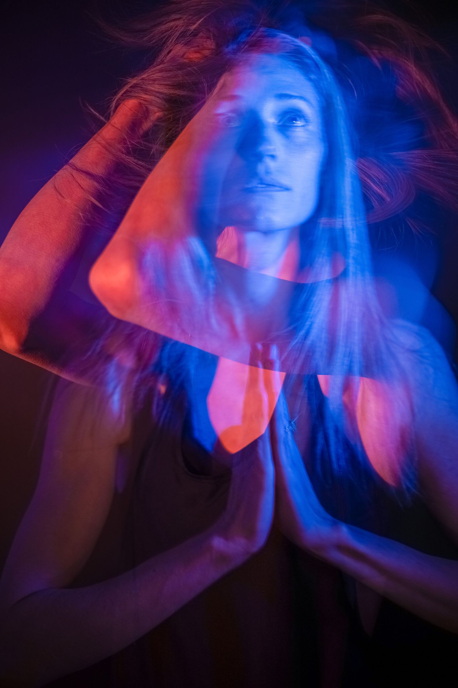 Artistic long exposure photo of woman with flowing hair illuminated by red and blue neon lights in darkness.