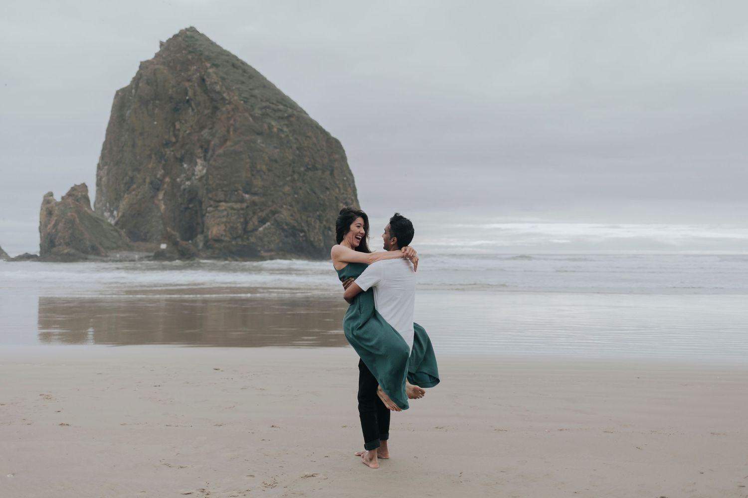Girl in teal smiling at bou while he picks her up and spins her on the beach in front of large rock in the ocean