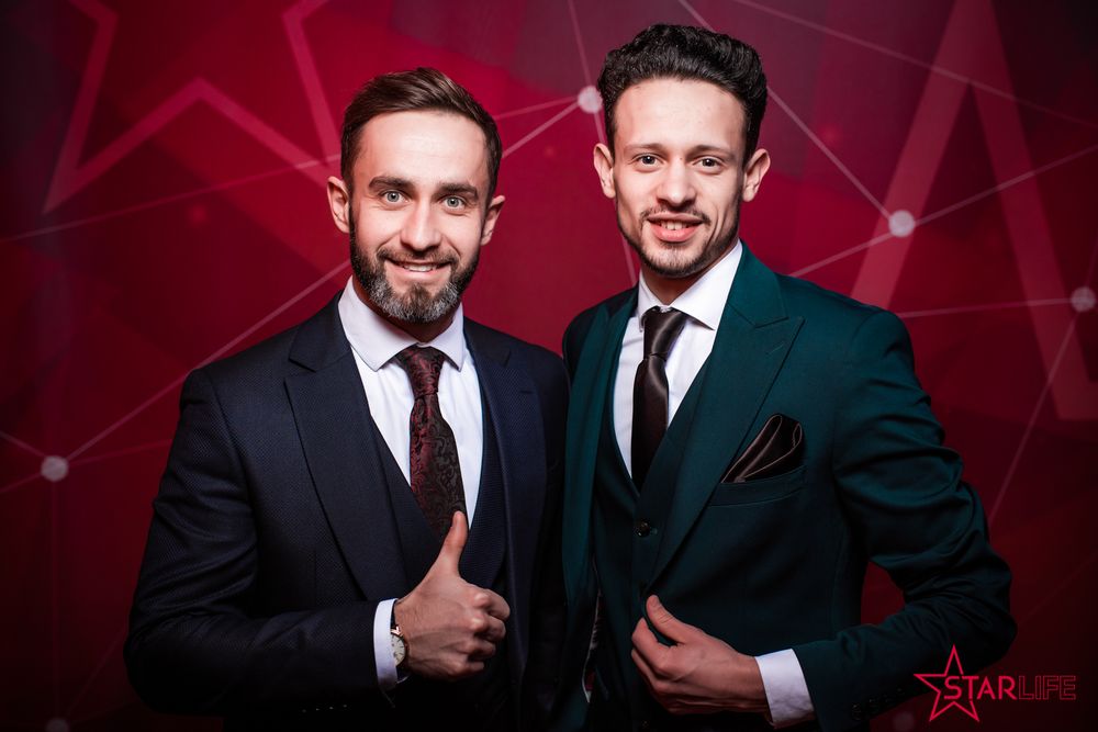 Two men in formal suits pose together against a red geometric background at an event.