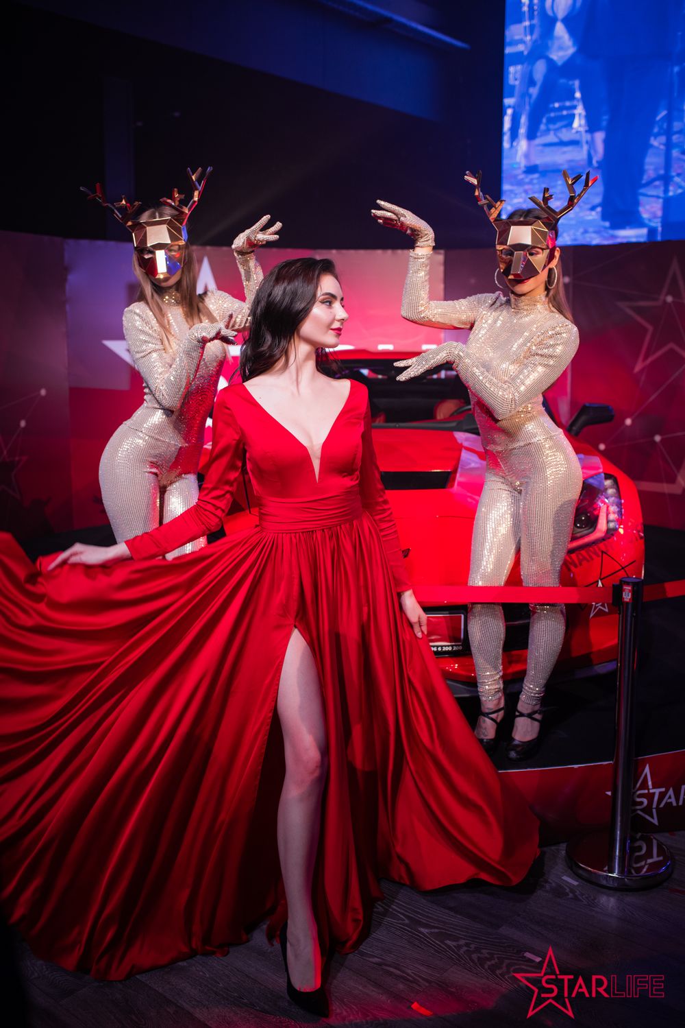 Woman in flowing red gown performs with dancers in reindeer costumes on stage.
