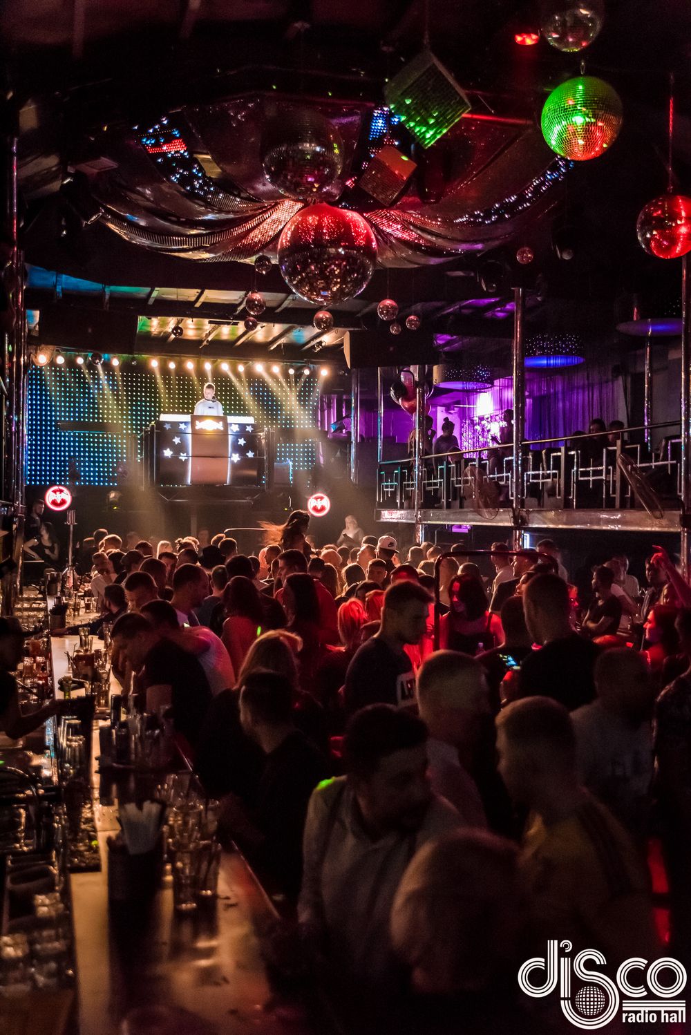 Packed nightclub interior with disco balls, colorful lighting and a large crowd enjoying the atmosphere.