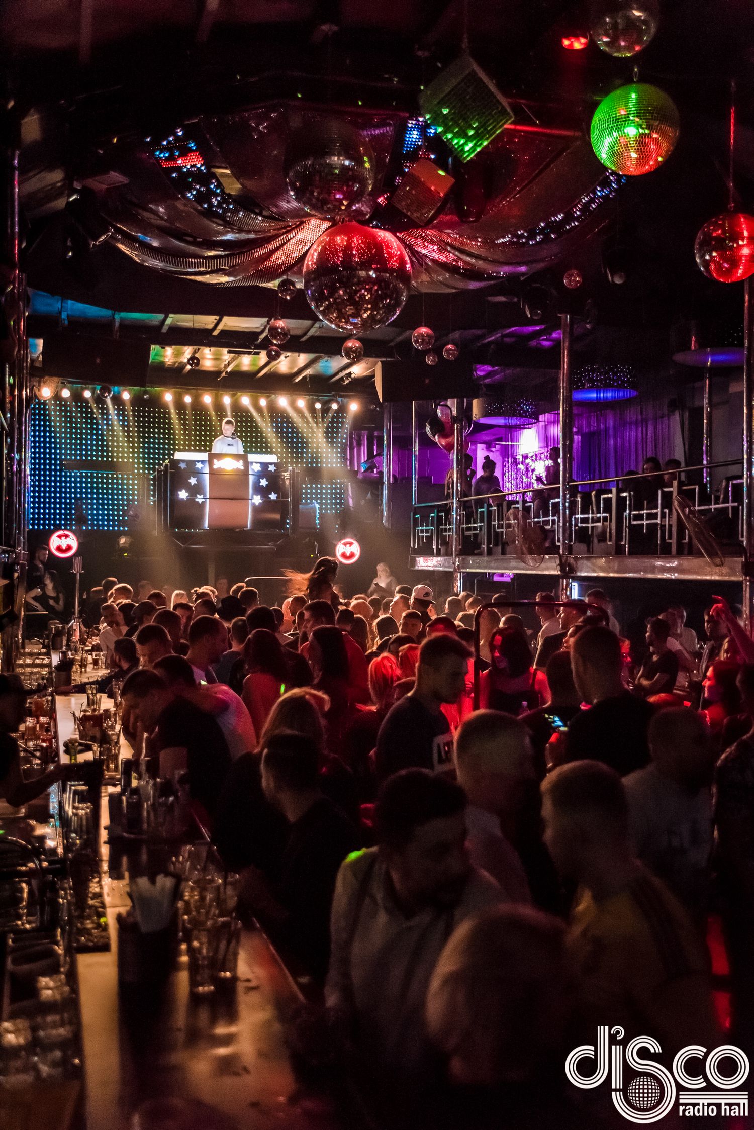 Packed nightclub interior with disco balls, colorful lighting and a large crowd enjoying the atmosphere.