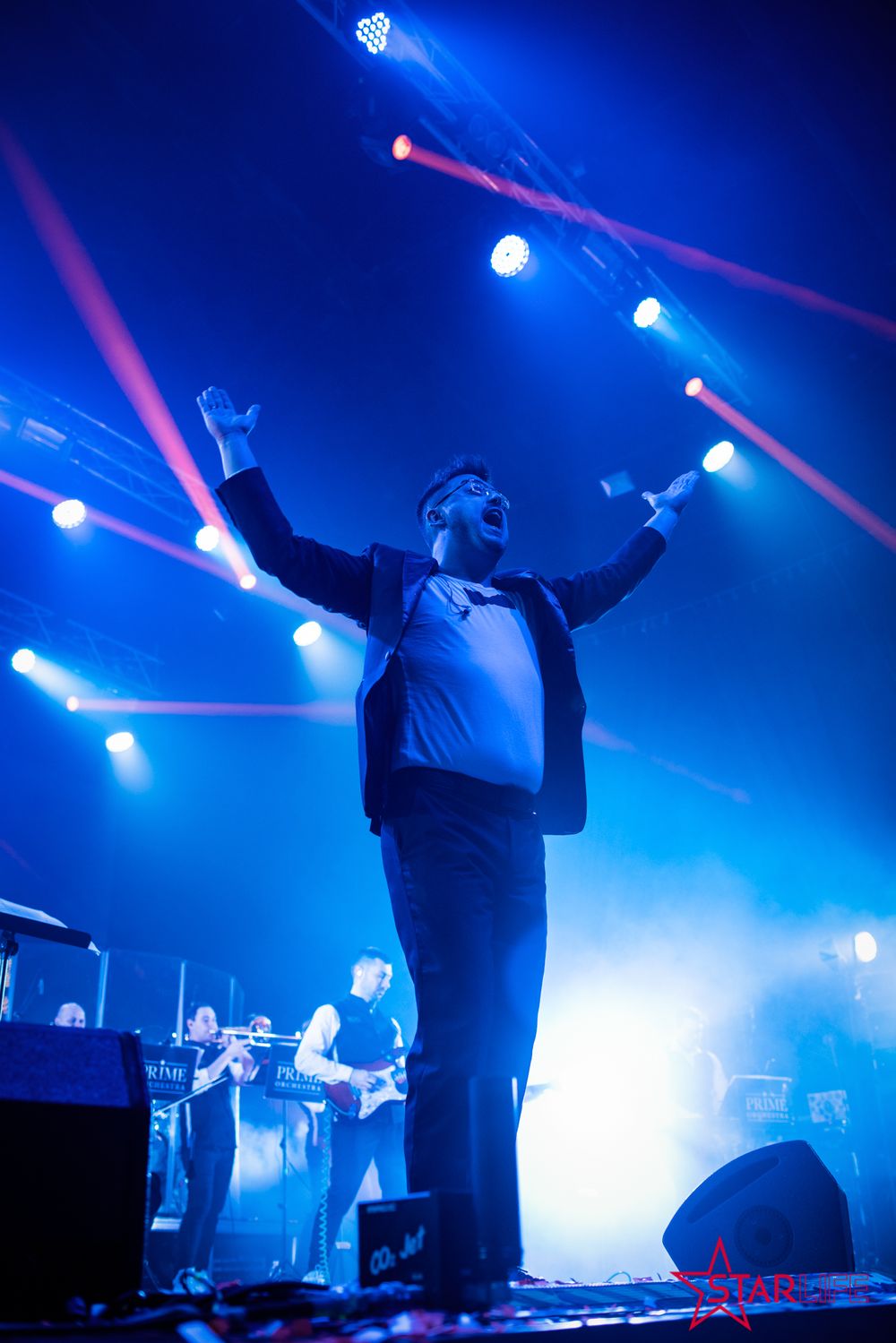 Performer on stage with arms raised surrounded by dramatic blue lighting and laser beams.