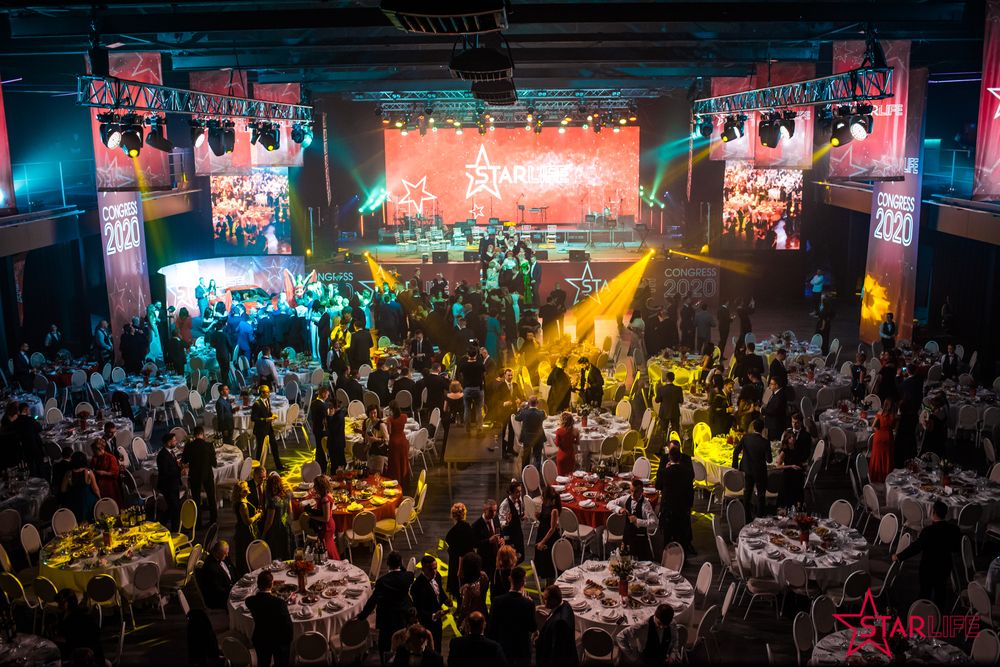 Large event venue with concert lighting and round tables filled with guests during a celebration.