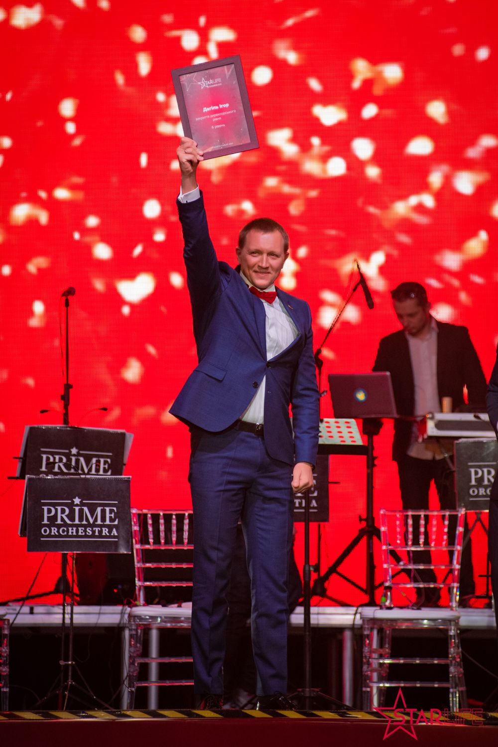 Performer in blue suit holds up award on stage with red backdrop and falling confetti effect.