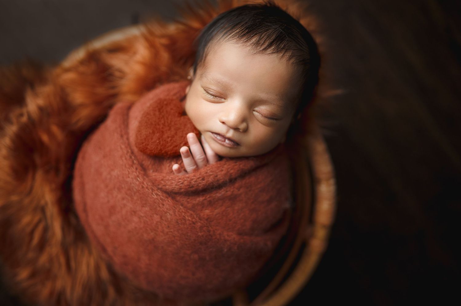 Serene newborn baby sleeping while wrapped in rust-colored swaddle on wooden surface.