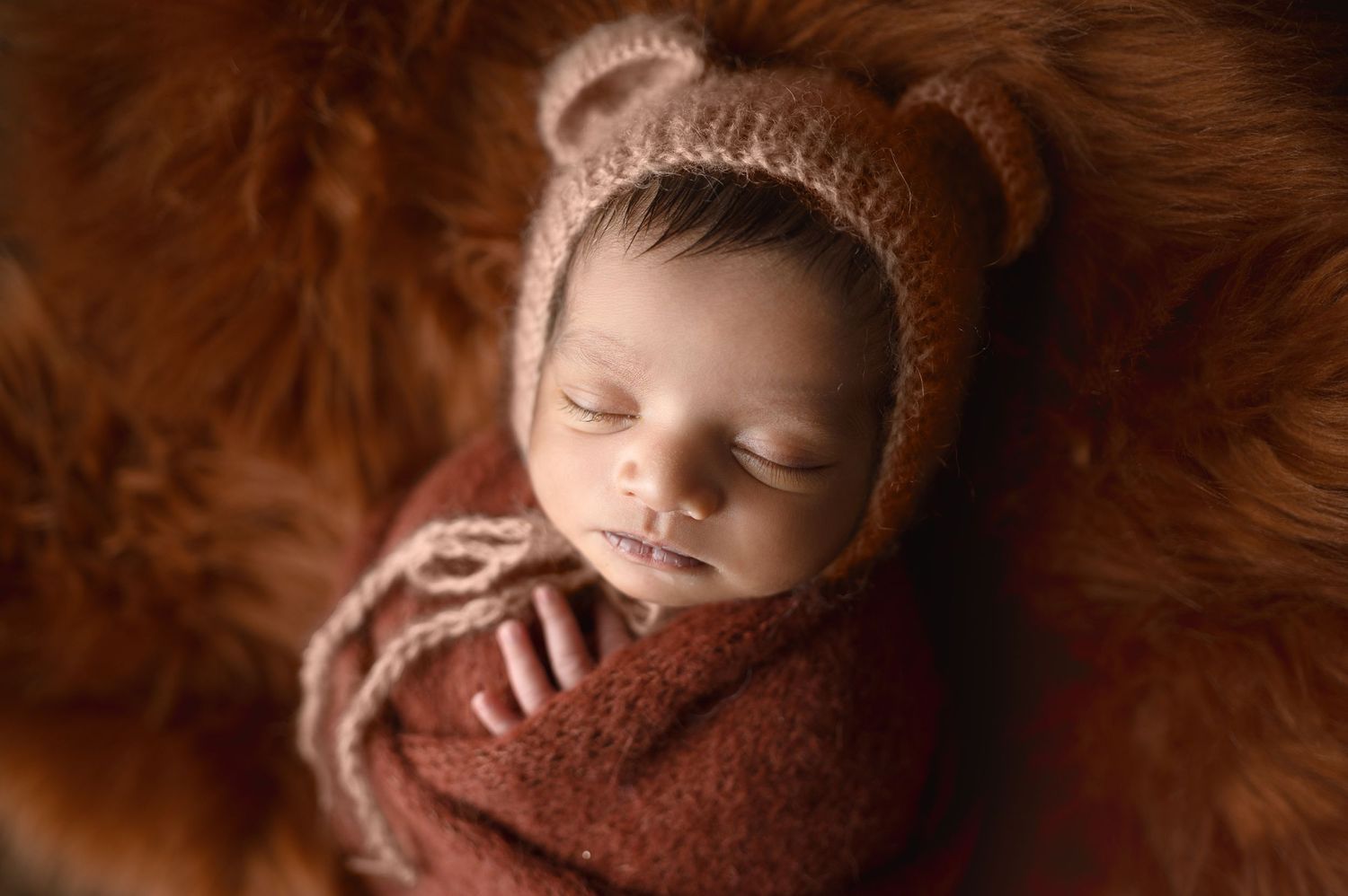 Sleeping newborn wearing brown knit bear bonnet swaddled in brown wrap.