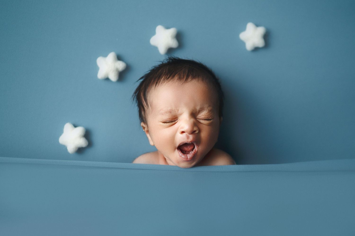 Baby yawning with mouth wide open against blue backdrop with white stars.