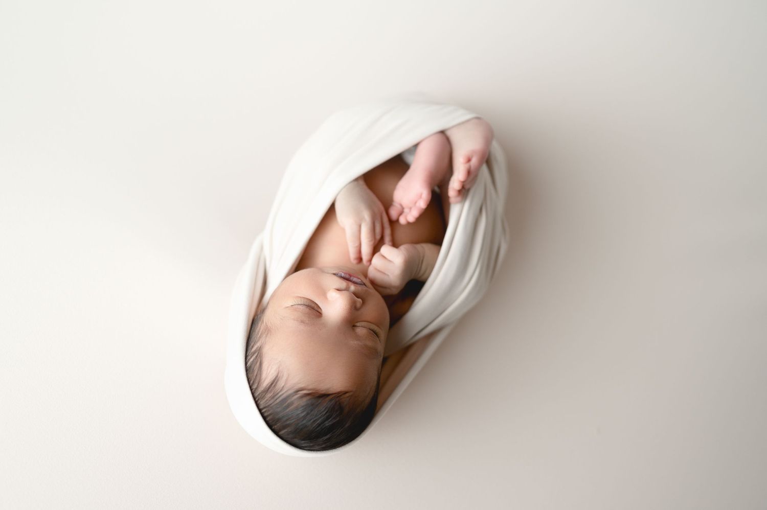 A sleeping newborn baby wrapped in white fabric during an artistic overhead portrait shoot.