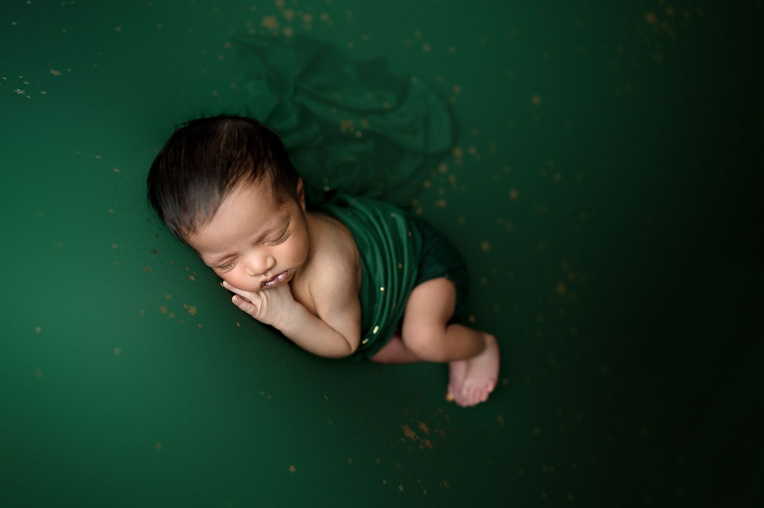 Peaceful sleeping newborn wearing green wrap against emerald backdrop with artistic lighting.