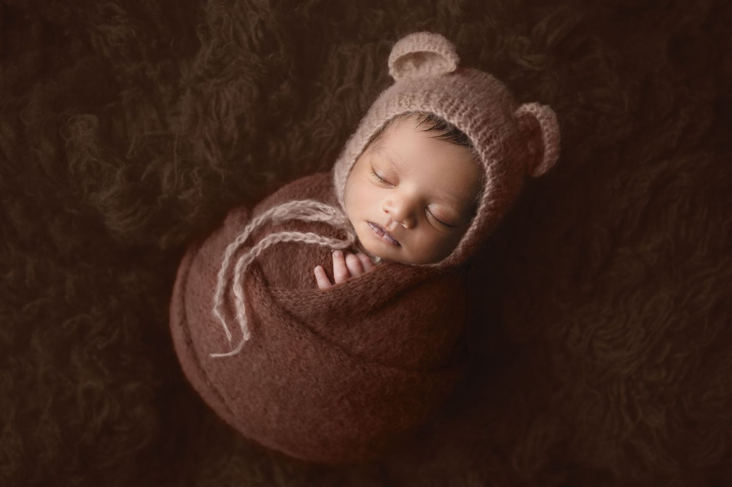 Newborn in bear hat swaddled in brown wrap on textured brown background.