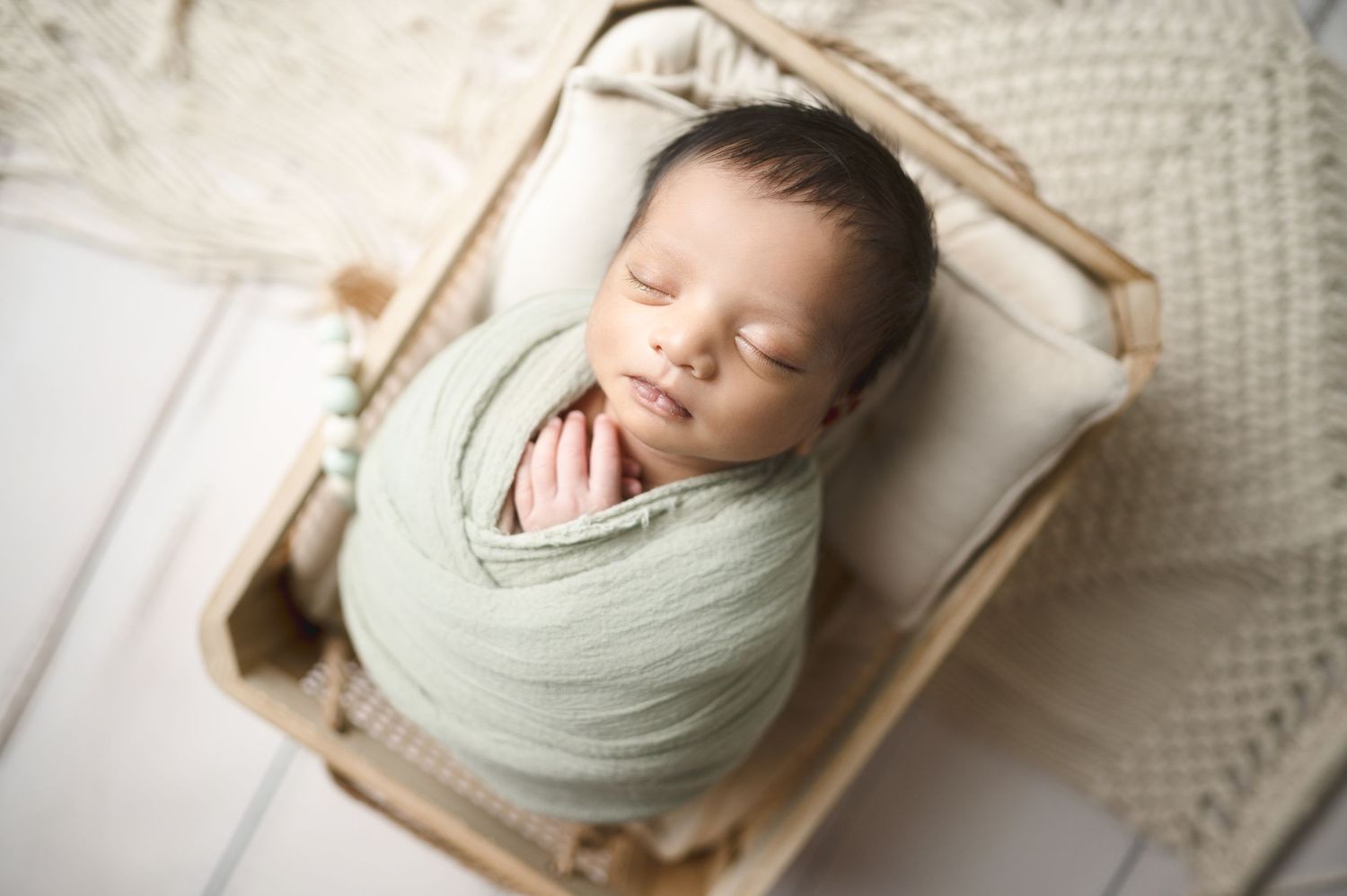 Sleeping newborn wrapped in soft mint green swaddle resting in wooden basket.