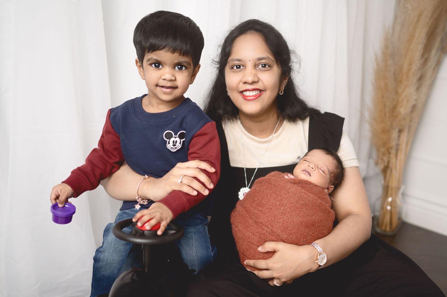 Family portrait with toddler and newborn in warm earth tones against white backdrop.