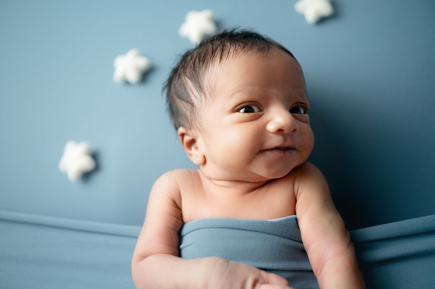 Alert newborn swaddled in blue fabric against backdrop with white stars.
