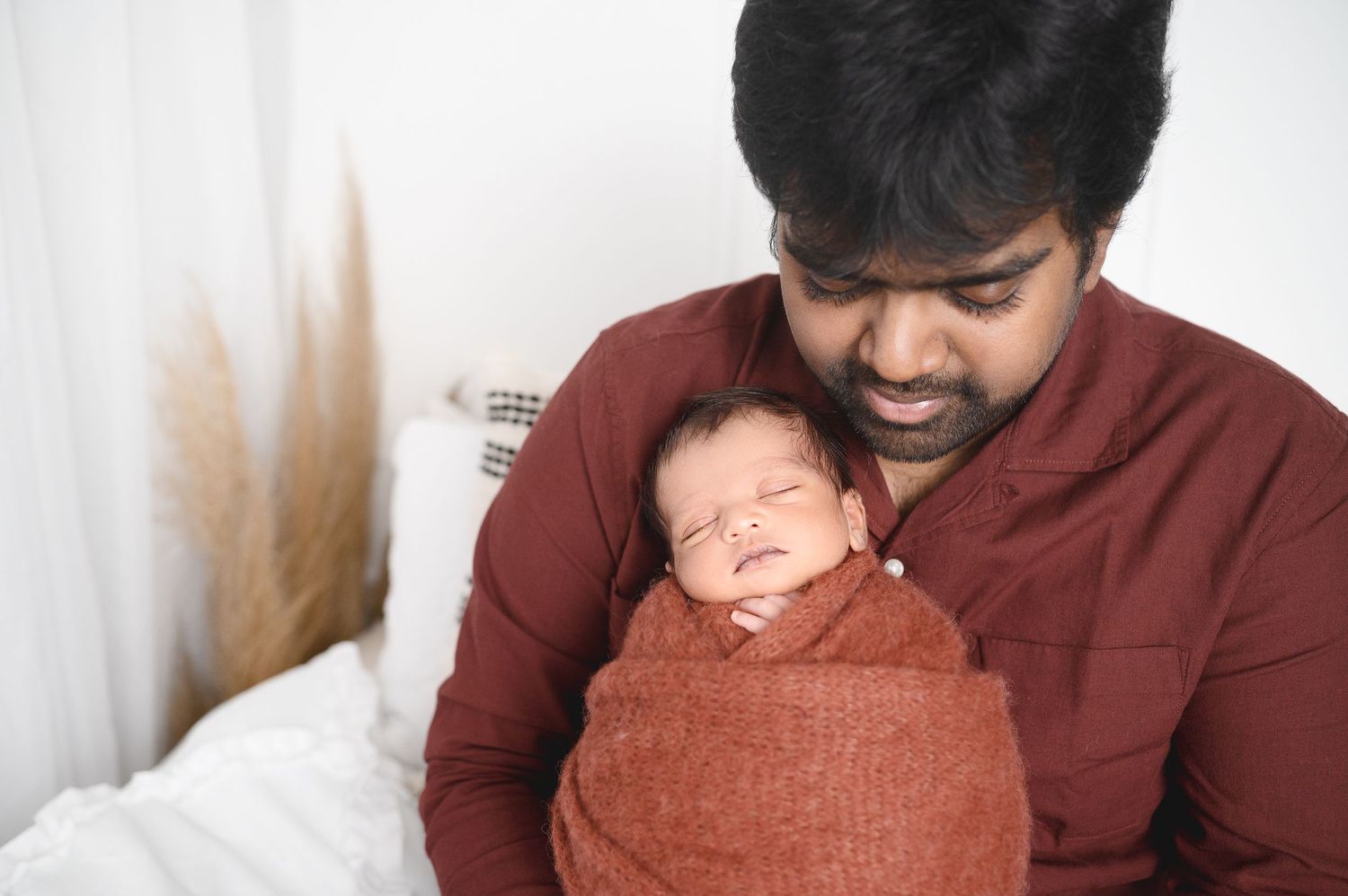 A tender moment between parent and sleeping newborn wrapped in rust-colored swaddle.
