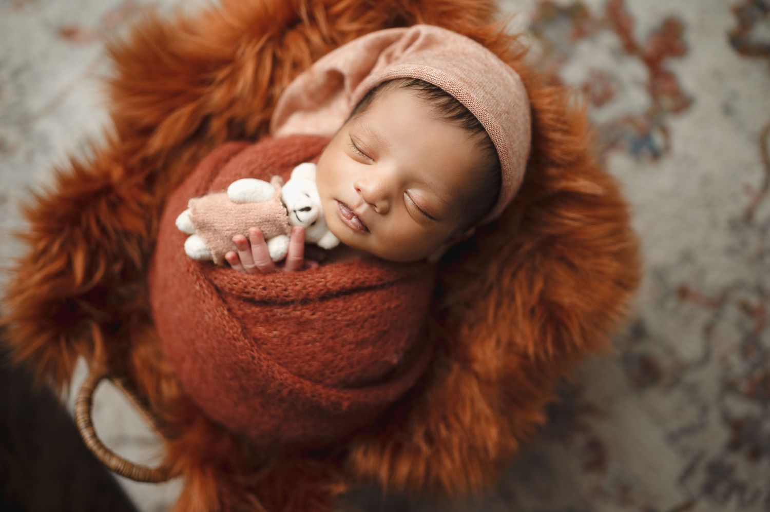 Sleeping newborn wearing beige cap and wrapped in rust swaddle on orange furry backdrop.