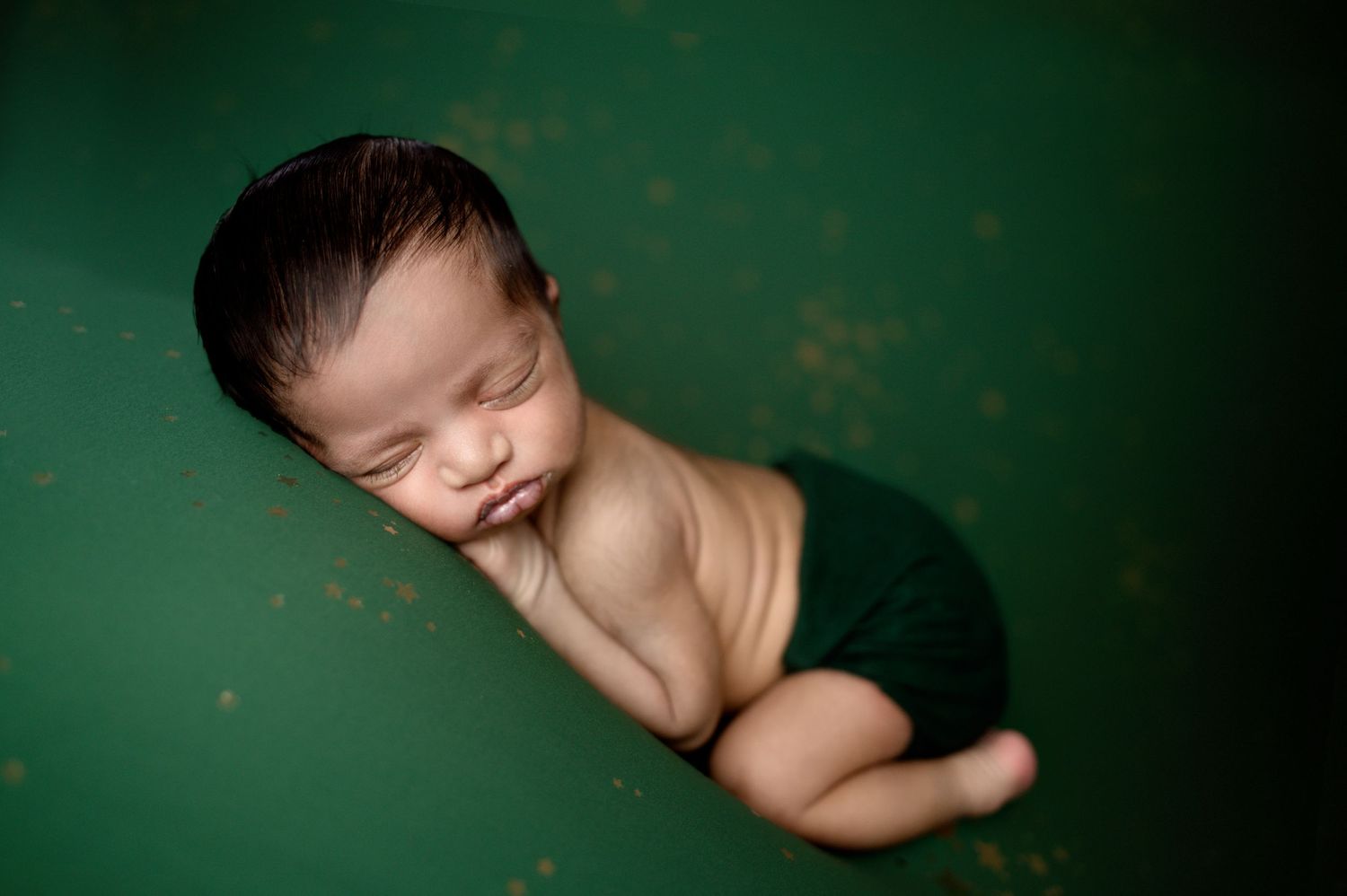Sleeping newborn baby posed on deep green background with artistic lighting.