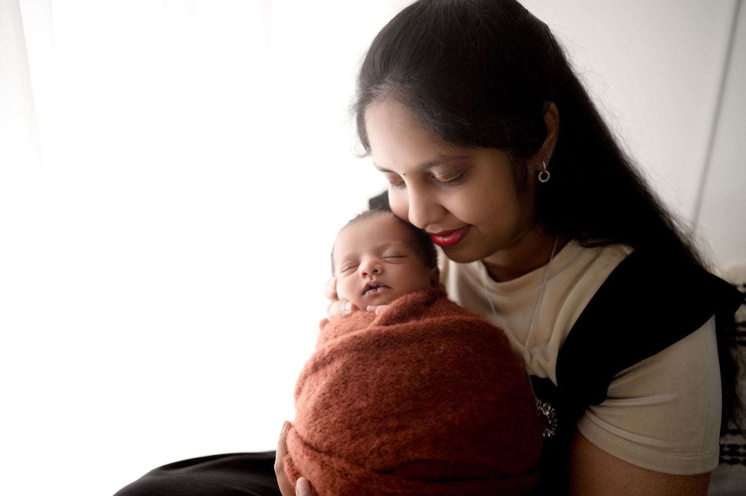 A tender moment captured between parent and newborn baby wrapped in rust-colored swaddle against light background.