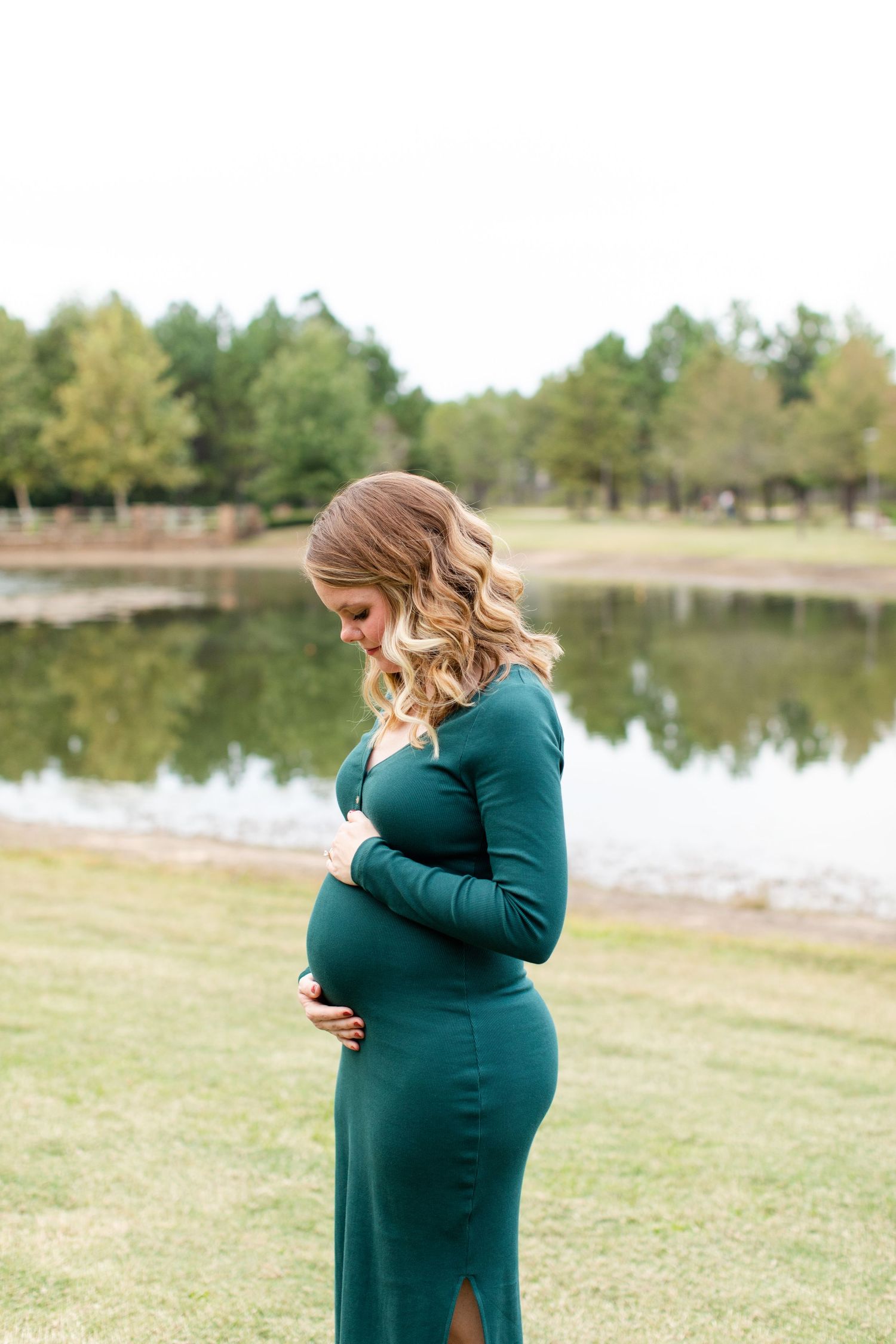 Expectant mother in teal dress poses by lakeside while cradling her baby bump.