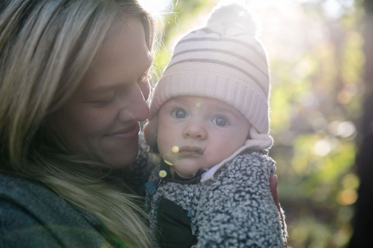 Sunlit outdoor portrait showing loving moment between caregiver and baby in winter clothing.