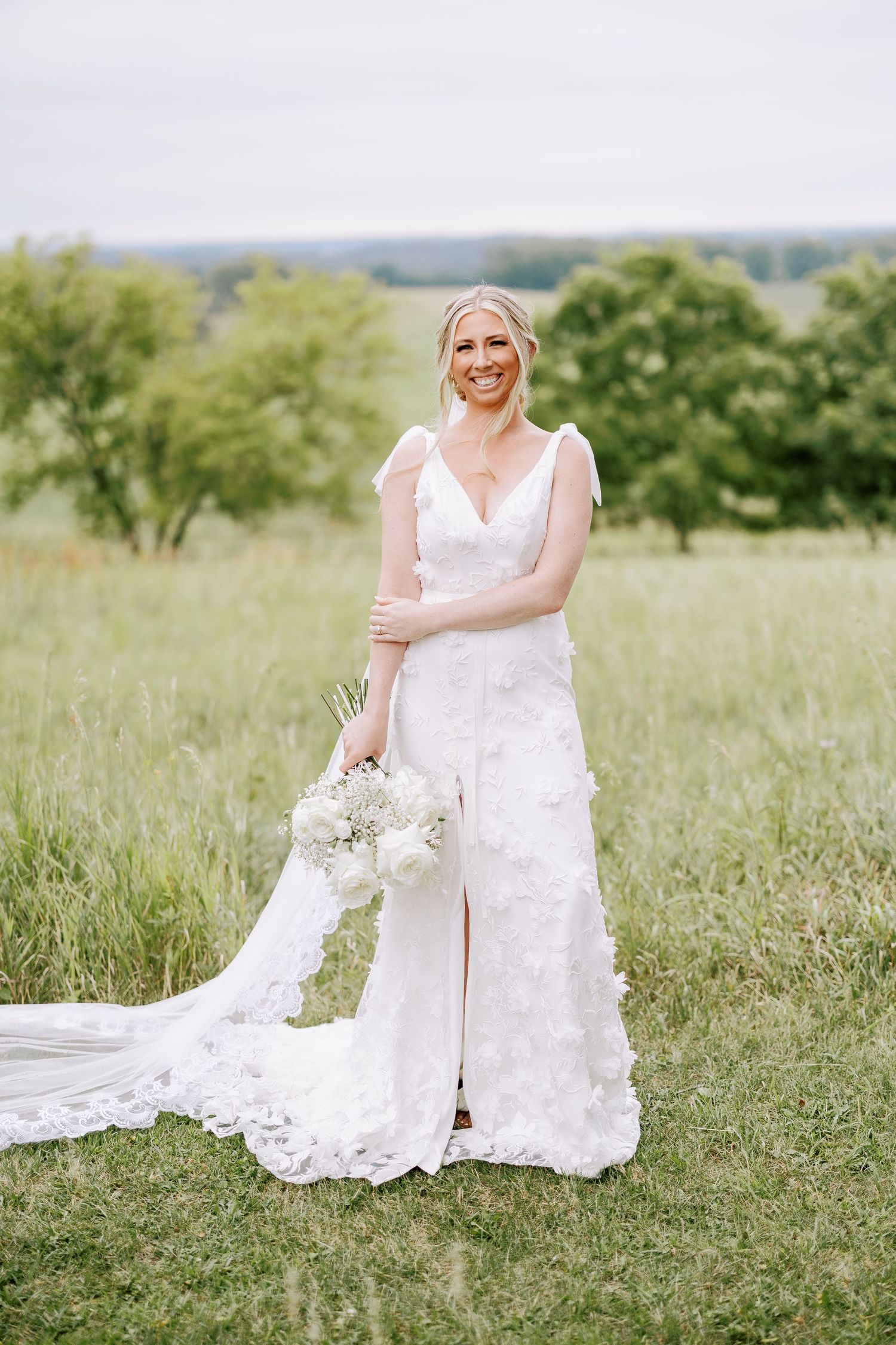 Bride in elegant white lace wedding gown poses in outdoor field setting.