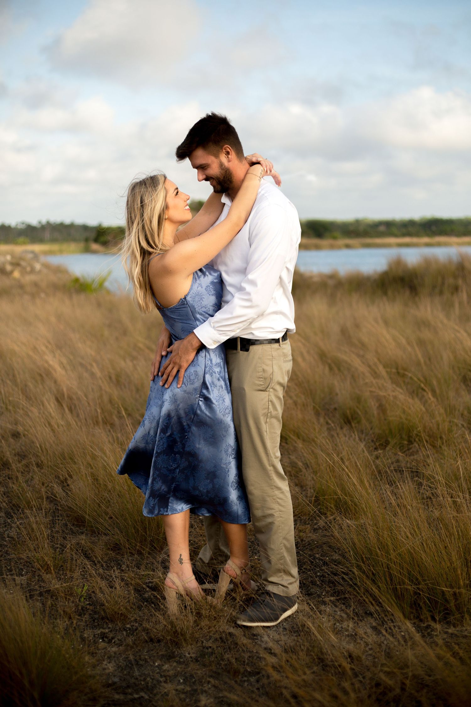 Romantic couple embracing in tall grass field with water view at sunset.