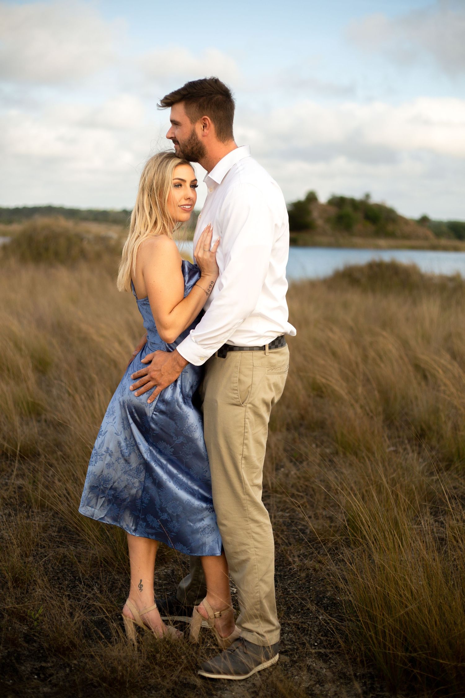 Loving couple poses together in blue dress and white shirt against coastal scenery.
