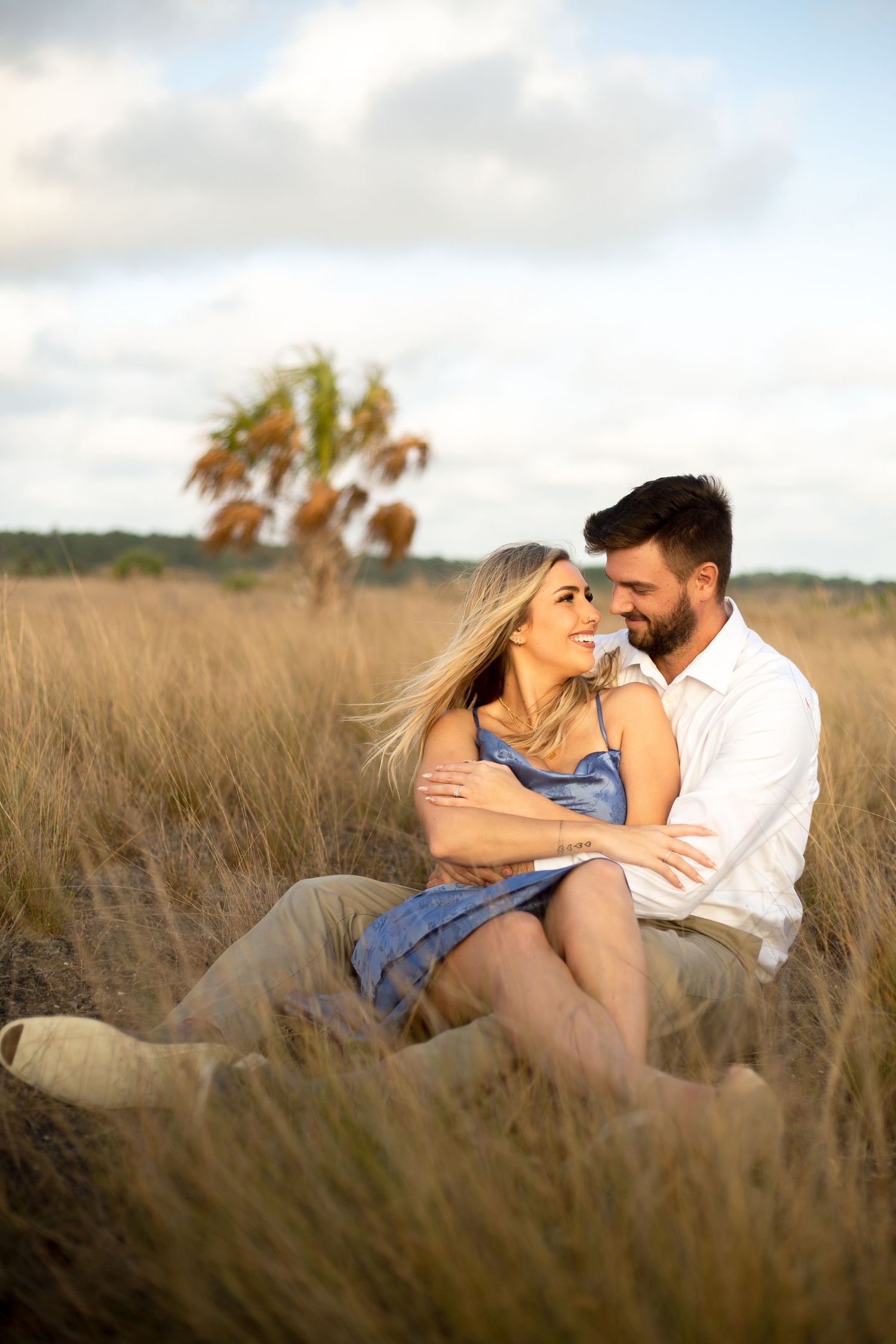 Loving couple cuddling in tall grass with palm trees silhouetted against cloudy sky.