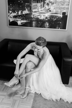 A figure in a white wedding dress fastens elegant heels while sitting on a dark couch beneath a cityscape photo.
