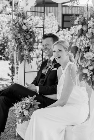 Couple sits on swing decorated with floral arrangements during outdoor ceremony.