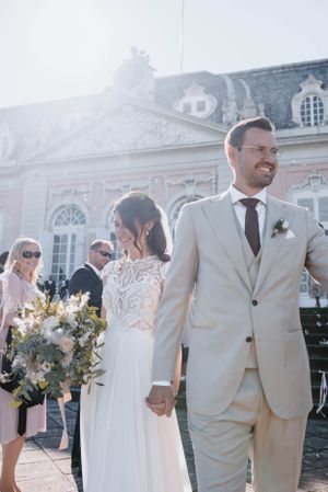 Elegant wedding couple walking hand in hand outside historic mansion in sunlight.
