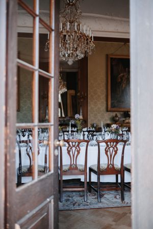 Elegant dining room with crystal chandelier viewed through doorway with antique wooden chairs.