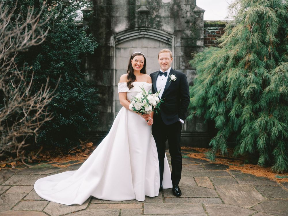 Newlyweds stand together before a stone architectural feature holding a white floral bouquet.
