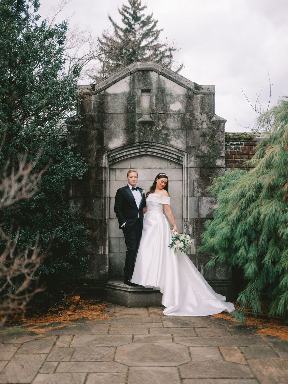 Wedding portrait featuring classic formal attire against weathered stone archway and greenery.