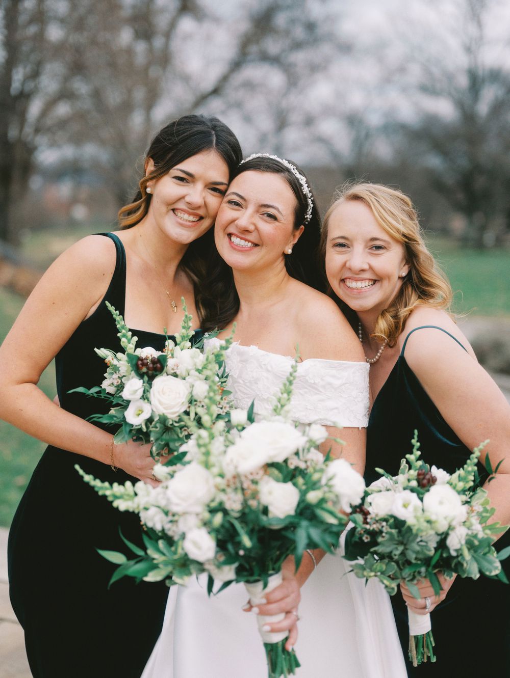 Bridesmaids in black dresses pose with white floral bouquets at an outdoor wedding.