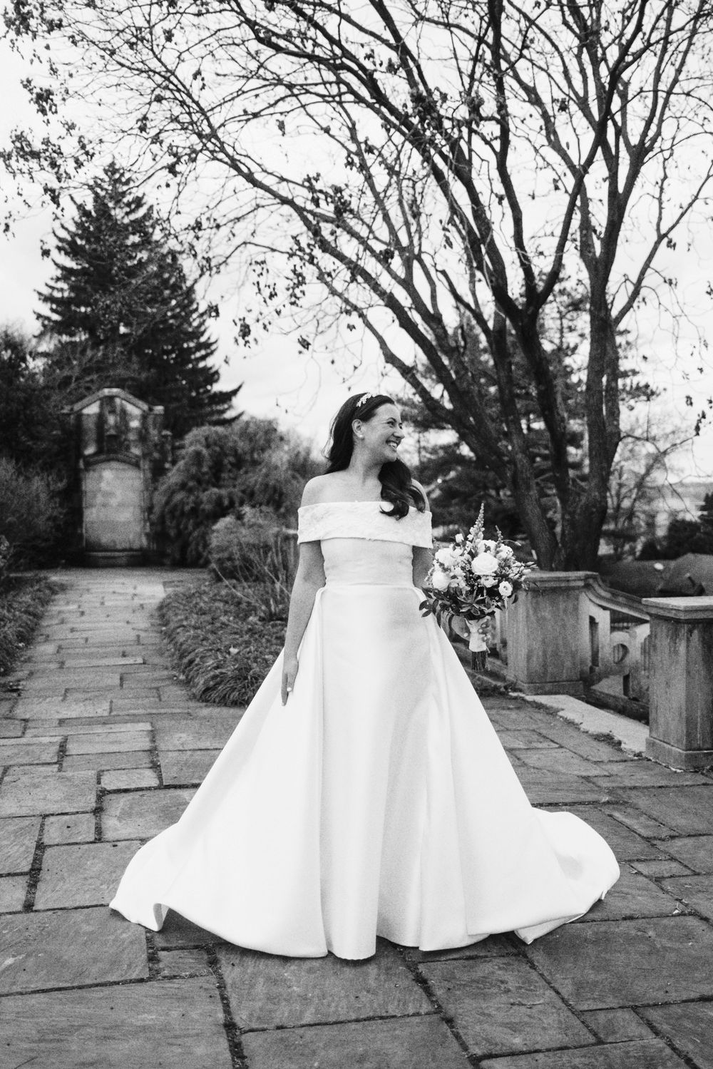 White wedding dress billowing in breeze on stone path surrounded by bare trees.