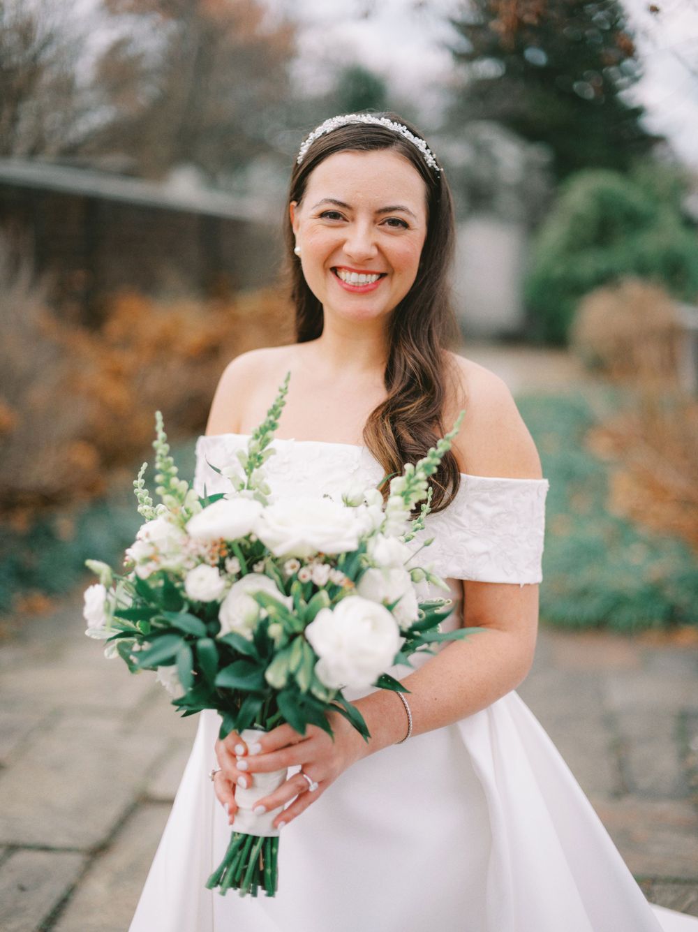 A bride in an off-shoulder wedding dress holds a lush white and green bouquet in a garden setting.