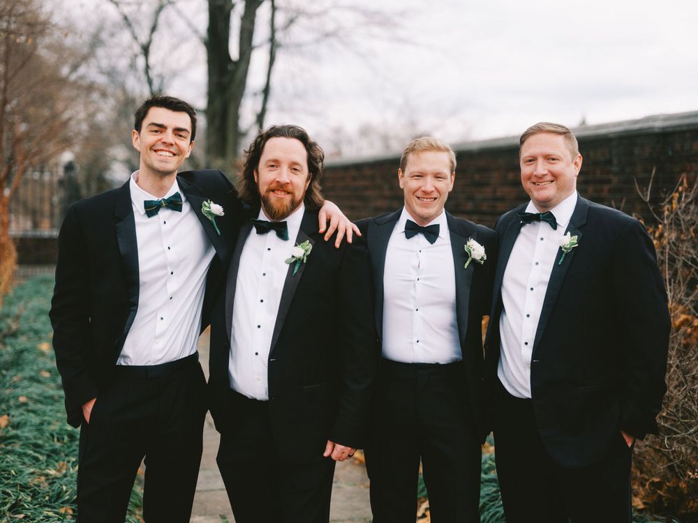 Four groomsmen in matching black tuxedos and bow ties pose together outdoors.