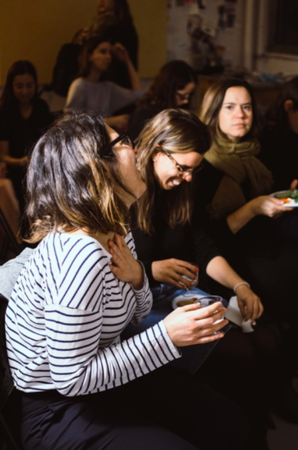 A group of friends sit together in casual conversation wearing cozy sweaters in a dimly lit room.