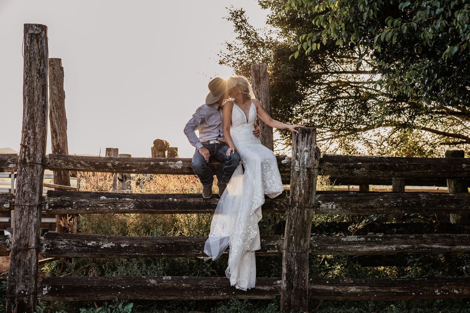 Couple poses for creative wedding photo on rustic wooden fence at sunset.