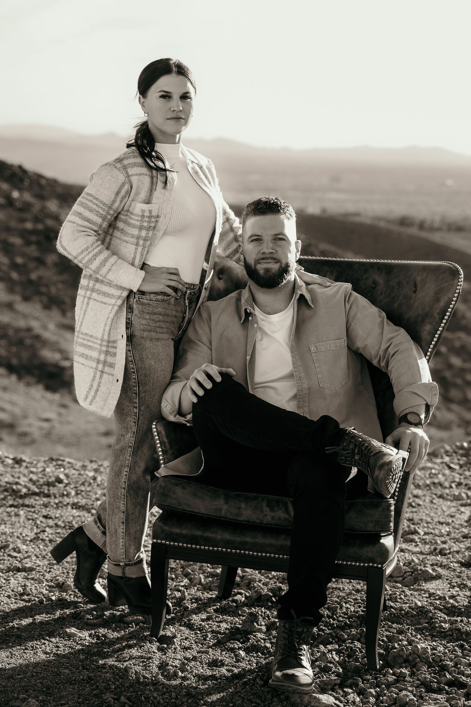 Stylish couple poses outdoors with leather armchair against scenic mountain backdrop in black and white photography.