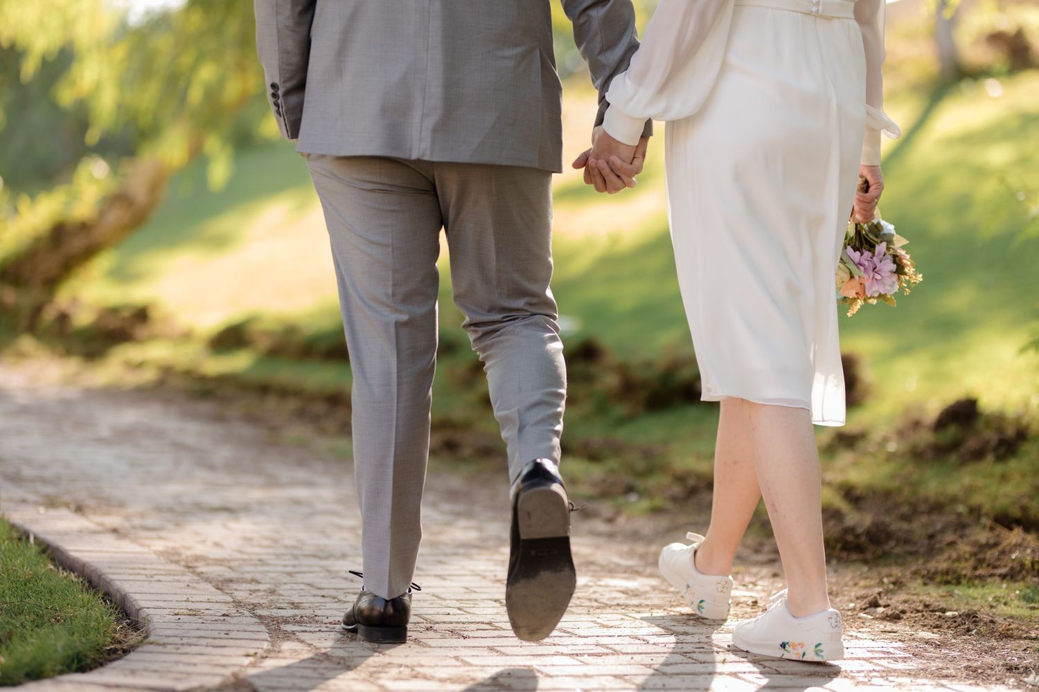Couple walking hand in hand on a garden path showing elegant formal attire from behind.