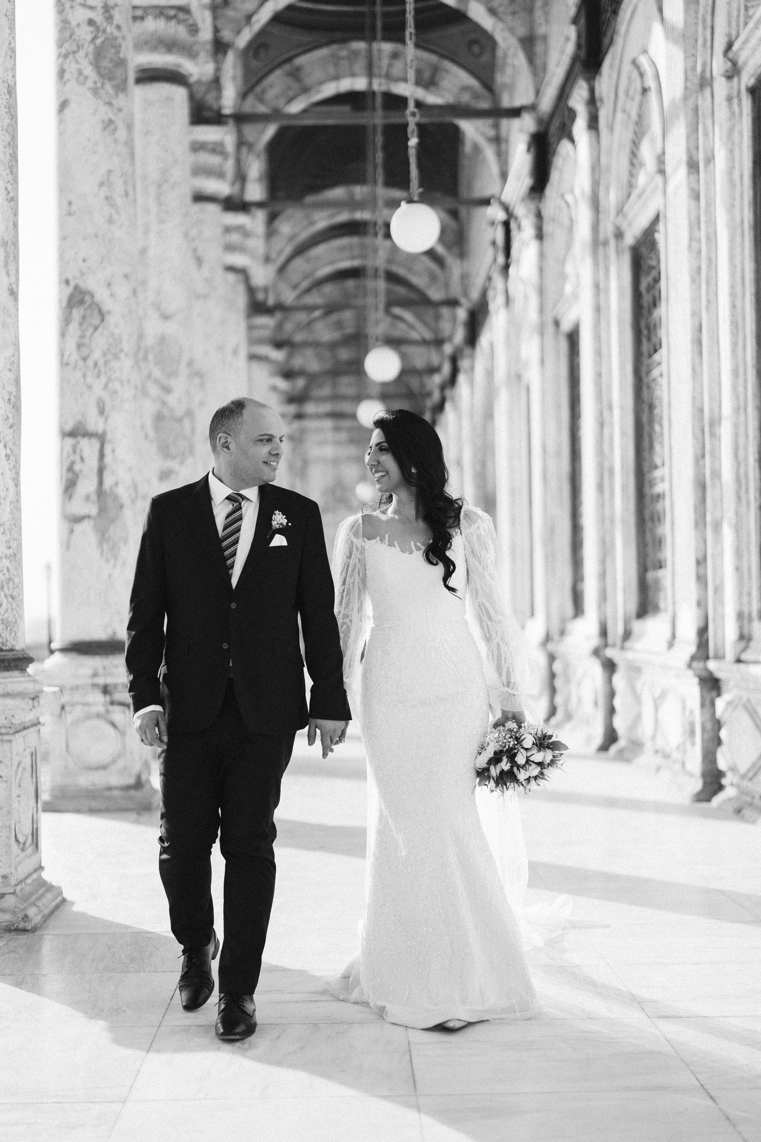 Elegant wedding couple walking through a historic colonnade with arched architecture.