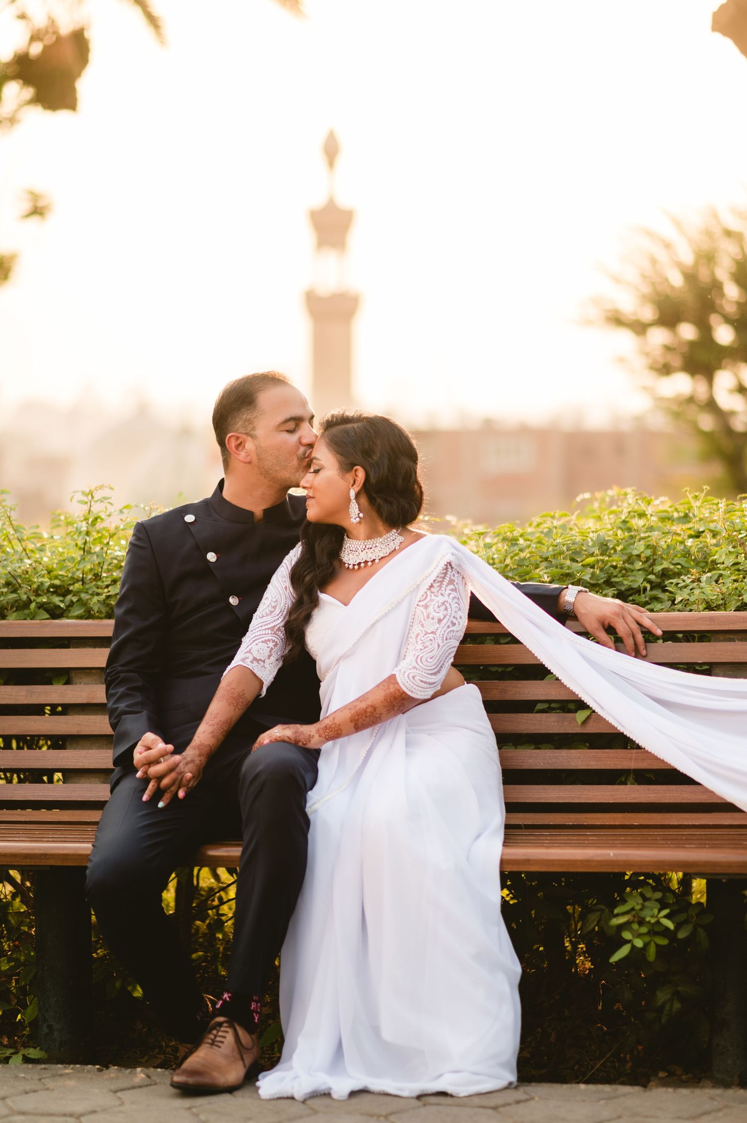 A tender wedding moment shared on a park bench with a mosque visible in the background.